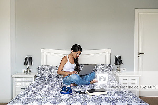 Lächelnde Frau im mittleren Erwachsenenalter  die einen Laptop benutzt  während sie mit einem Buch und Kopfhörern auf dem Bett zu Hause sitzt