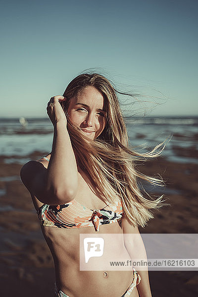 Lächelnde junge Frau im Bikini steht gegen das Meer und den klaren Himmel bei Sonnenuntergang