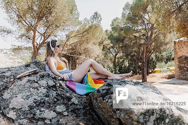 Junge Frau mit Sonnenbrille und Regenbogenflagge auf einem Felsen sitzend