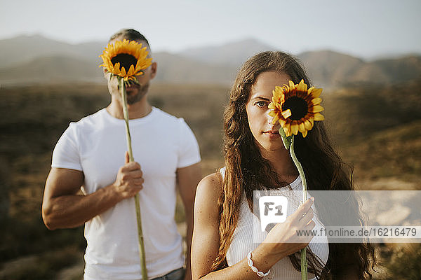 Woman and man hiding face with sunflowers during sunset