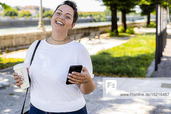 Porträt einer glücklichen  kurvigen jungen Frau mit Getränk zum Mitnehmen und Mobiltelefon in der Stadt