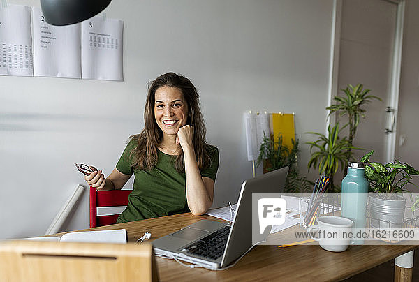 Lächelnde Geschäftsfrau sitzt an einem Schreibtisch an der Wand im Büro zu Hause