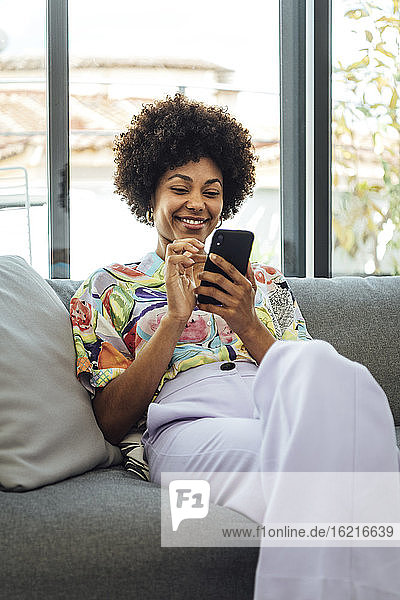 Glückliche Frau im mittleren Erwachsenenalter  die ein Smartphone benutzt  während sie auf dem Sofa in einem modernen Penthouse sitzt