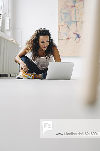 Fröhliche Frau im mittleren Erwachsenenalter  die einen Laptop benutzt  während sie zu Hause auf dem Boden sitzt