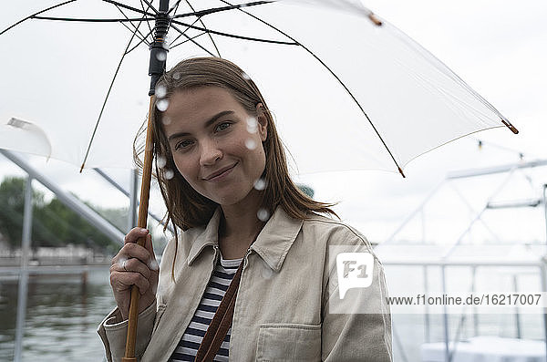 Nahaufnahme einer lächelnden jungen Frau mit Regenschirm  die bei Regen in der Stadt steht