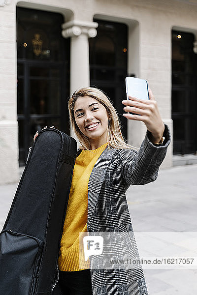 Young woman with violin case taking selfie in city