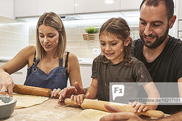 Lächelndes Mädchen mit Eltern knetet Pizzateig auf einem Tisch in der Küche