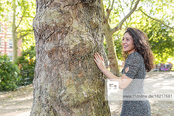 Fröhliche junge Frau schaut weg  während sie an einem Baumstamm im Park steht