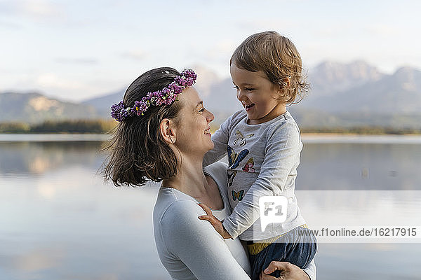 Lächelnde Mutter mit Diadem  die ihre Tochter am Ufer eines Sees trägt