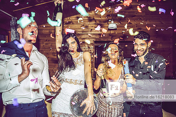 Männliche und weibliche Freunde mit Champagner und Discokugel tanzen inmitten von Konfetti in der Partei