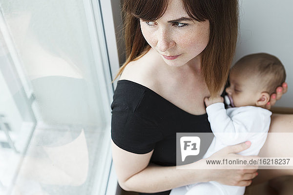 Junge Mutter mit ihrem kleinen Mädchen schaut zu Hause aus dem Fenster