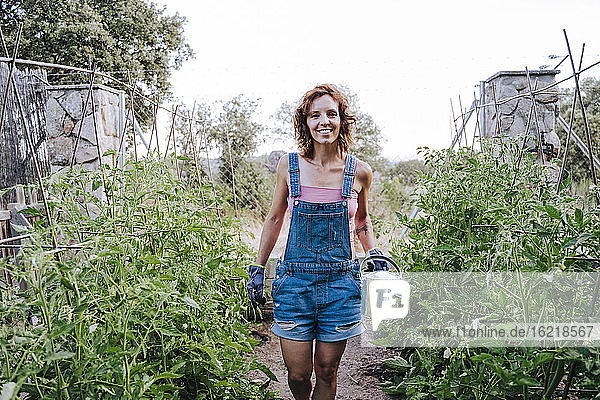 Lächelnde Frau  die eine Gießkanne hält  während sie inmitten von Pflanzen im Gemüsegarten steht
