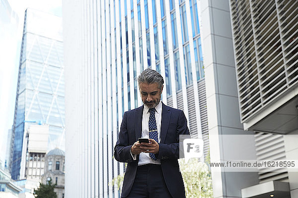 Geschäftsmann  der vor einem Bürogebäude in der Stadt eine SMS mit seinem Mobiltelefon schreibt