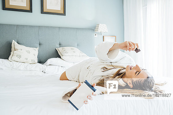Frau hält Smartphone und isst Schokolade  während sie auf dem Bett im Hotel liegt