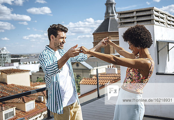 Glückliches multiethnisches Paar tanzt auf der Penthouse-Terrasse an einem sonnigen Tag