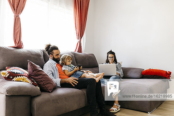 Frau arbeitet am Laptop  während Vater und Tochter auf dem Sofa zu Hause ein Smartphone benutzen