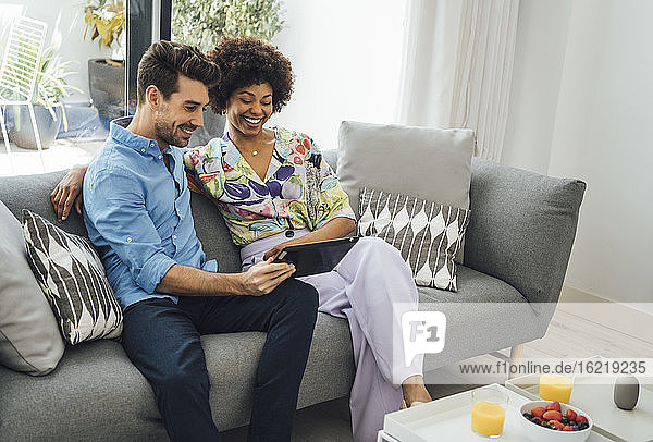 Ein glückliches Paar benutzt ein digitales Tablet  während es auf dem Sofa im Wohnzimmer eines Penthouses sitzt.