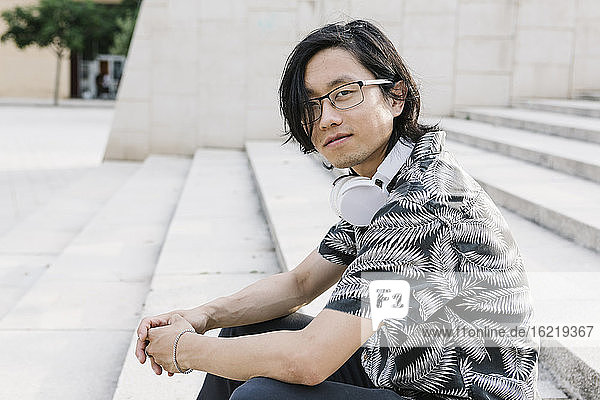 Young man wearing eyeglasses with headphones sitting on steps in city