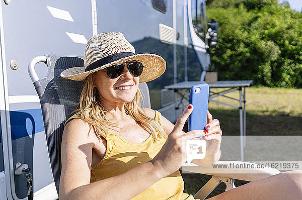 Relaxed woman sitting next to camper enjoying vacation and using smartphone
