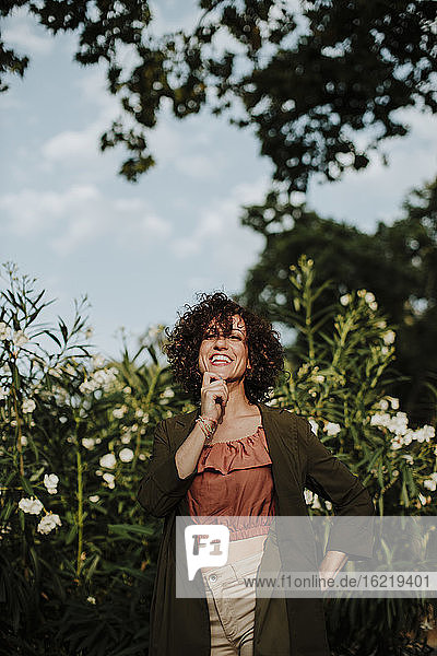 Lächelnde mittelgroße erwachsene Frau mit lockigem Haar und Mantel  die gegen Pflanzen im Park steht