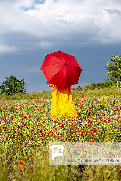 Ältere Frau mit rotem Regenschirm in einem Mohnfeld gegen den Himmel stehend