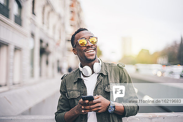 Cheerful man wearing sunglasses using smart phone while standing in city