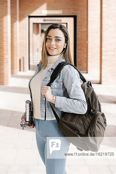 Smiling student with bag and book standing in university campus