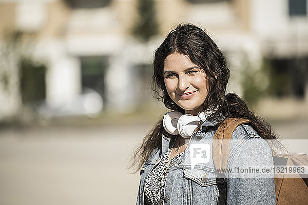 Lächelnde Studentin mit Rucksack und Kopfhörern in der Stadt an einem sonnigen Tag