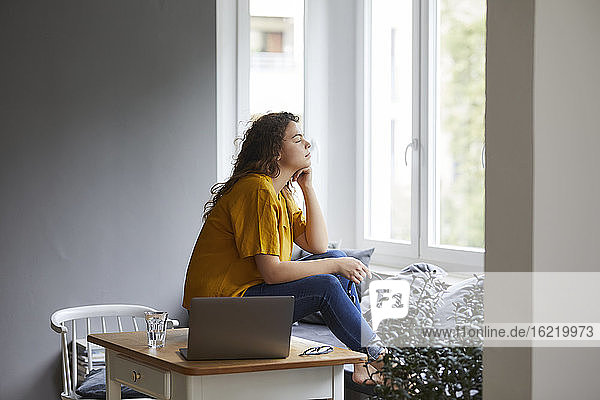 Nachdenkliche Frau mit Hand am Kinn  die durch das Fenster schaut  während sie eine Pause von der Arbeit im Wohnzimmer im Büro zu Hause macht