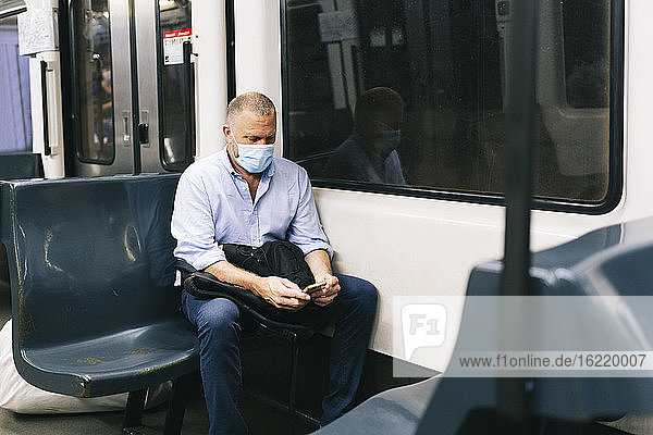 Geschäftsmann mit Gesichtsmaske  der während der Fahrt im Zug sein Smartphone benutzt