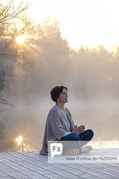 Erwachsene Frau meditiert auf einem Steg am Seeufer bei nebligem Sonnenaufgang