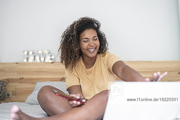 Fröhliche junge Frau mit Smartphone und Laptop auf dem Bett zu Hause