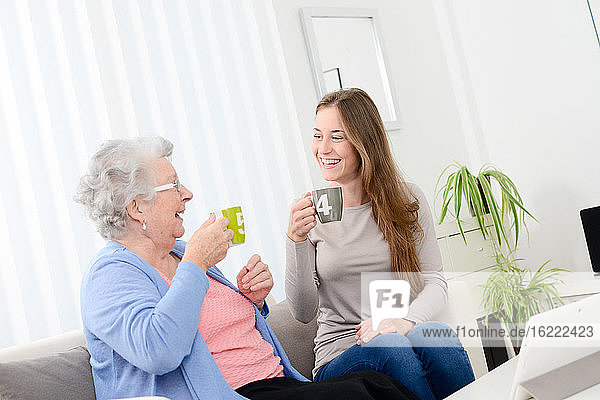 Happy old senior woman spending time drinking tea with cheerful young girl at home