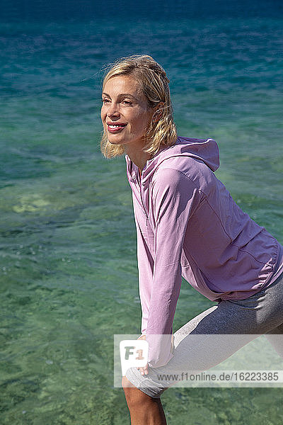 Eine schöne lächelnde junge Frau in Sportkleidung  die sich vor dem Meer in der Sonne räkelt.
