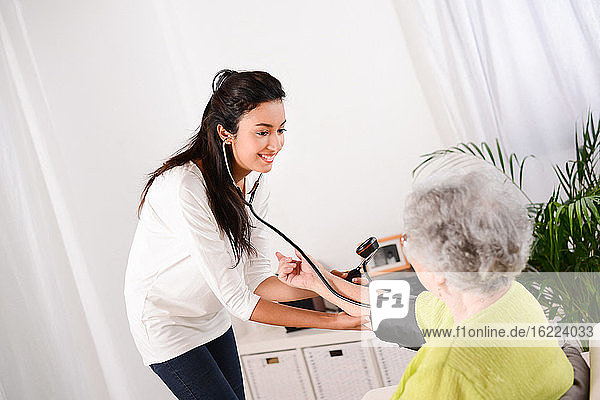Fröhliche junge Ärztin  die bei einer älteren Frau zu Hause den Blutdruck misst