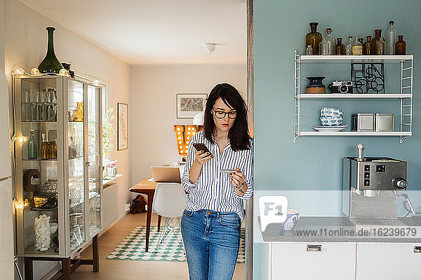 Young woman with smartphone and credit card in kitchen
