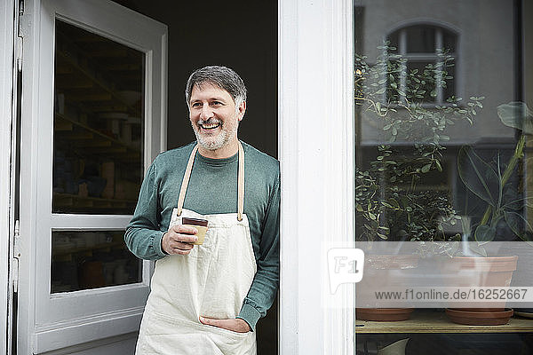 Lächelnder reifer männlicher Besitzer schaut weg  während er beim Kaffee an der Tür des Kunstateliers einen Kaffee trinkt