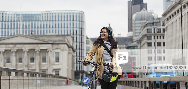 Lächelnde Geschäftsfrau mit Kopfhörern beim Radfahren auf der Stadtbrücke