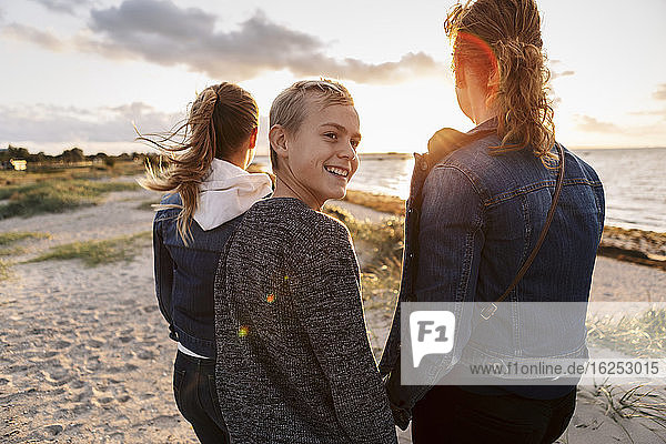 Smiling teenager looking away while standing with mother and daughter at beach