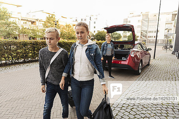 Siblings with suitcase walking while smiling mother standing by car on road
