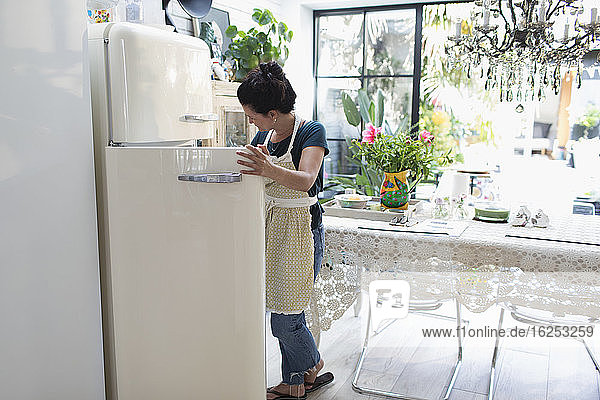 Woman in apron standing at open refrigerator in kitchen