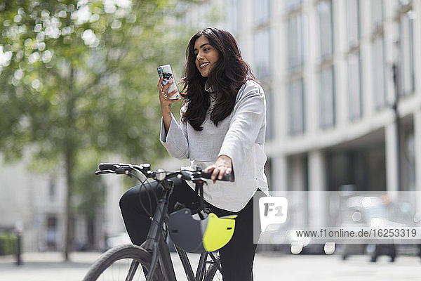 Woman on bicycle using smart phone on sunny city street