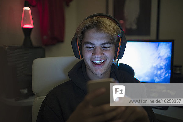 Lächelnder Teenager mit Kopfhörern  der ein Smartphone in einem dunklen Raum benutzt