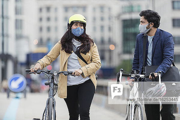 Business people in face masks walking bicycles on city bridge