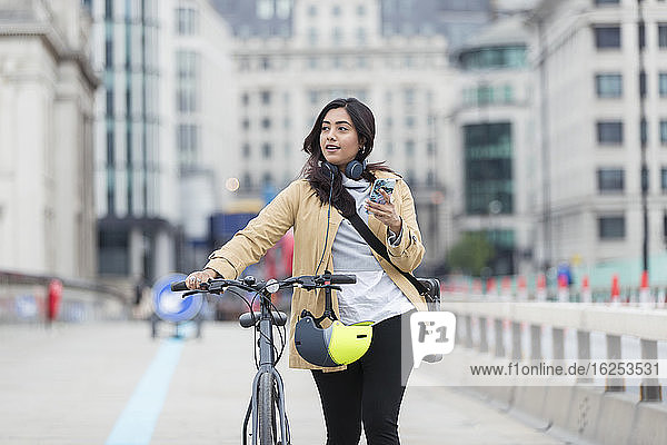 Frau mit Smartphone geht Fahrrad auf Stadtbrücke