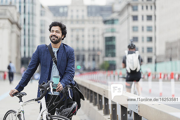 Smiling businessman with bicycle and coffee on city bridge