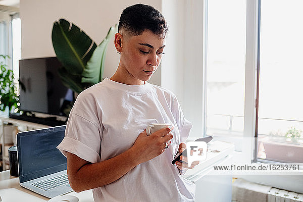 Young woman with shaved head standing in a kitchen  checking her mobile phone.