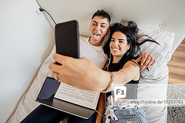Junges lesbisches Paar liegt auf einem Bett und macht ein Selfie mit dem Handy.