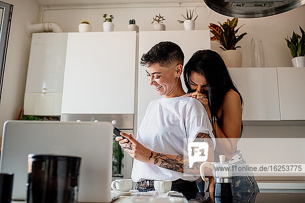 Junges lesbisches Paar steht in der Küche und schaut auf das Handy.