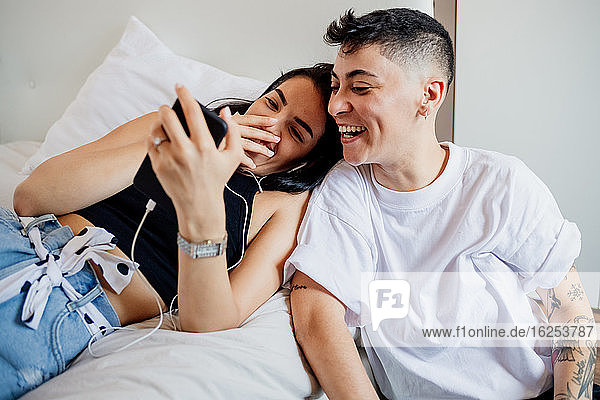 Junges lesbisches Paar in einem Schlafzimmer  schaut auf ein Handy  lächelt.